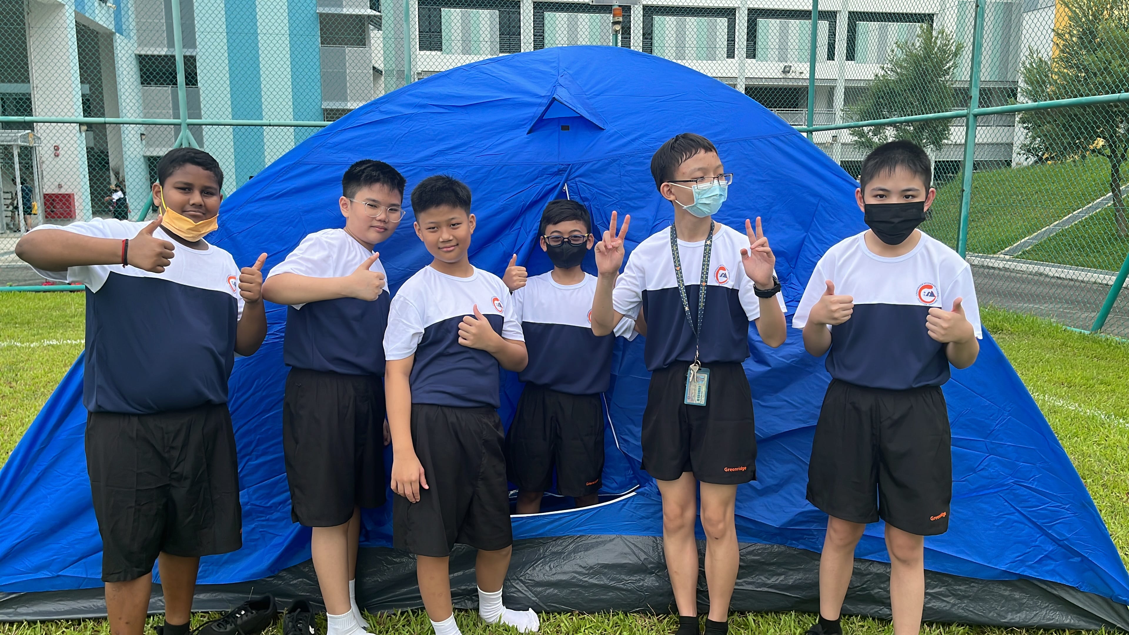 Six school boys in matching uniforms pose with thumbs up in front of a large blue dome tent outdoors.